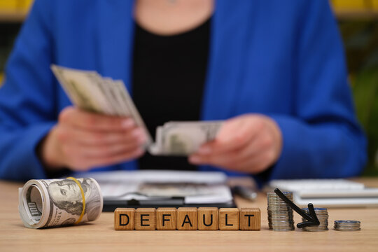 A person in a blue blazer counts cash while sitting at a desk. Blocks spelling default sit nearby, creating a sense of financial tension in a contemporary office filled with paperwork.