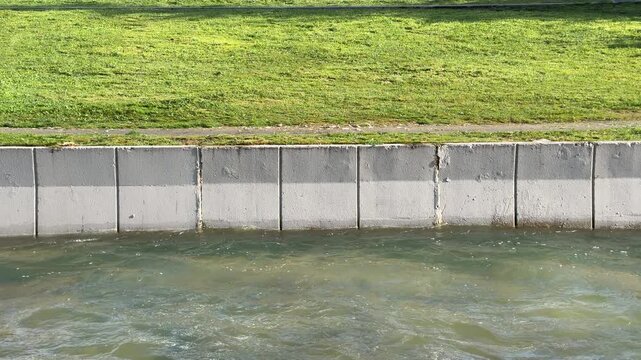 A sturdy concrete barrier separates a fast-flowing river from a bright green lawn. The scene feels structured and calm. Sunlight reflects off the moving water against the solid gray wall.