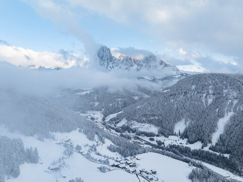 Aerial view of Chiesa di San Giacomo surrounded by snow-covered pine forests and traditional alpine houses with the Langkofel mountain peaks in the background Ortisei, Trentino-Alto Adige, Italy.