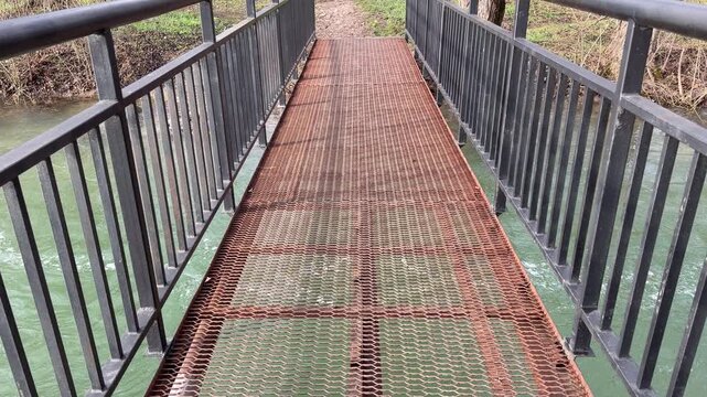 A weathered metal bridge with a rusty grate floor stretches across moving green water. The sturdy black railings offer a sense of safety against the wild, flowing river below.