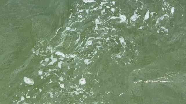 Churning green water flows steadily, creating soft ripples and trails of white foam. The murky depth evokes a sense of mystery and the powerful, relentless movement of nature.