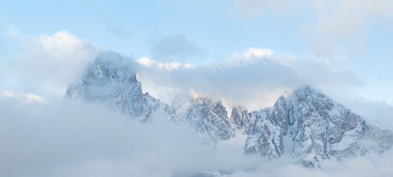 Aerial view of snow-capped mountain peaks partially hidden by thick white clouds under a clear blue sky in Ortisei, Trentino-Alto Adige, Italy.