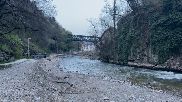A riverside path follows fast flowing water beneath a steel truss bridge in Uzice, Serbia, with rocky banks, steep ivy covered cliffs and bare trees.