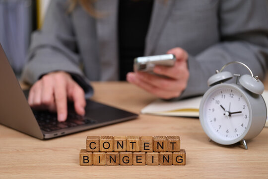 In a cozy workspace, someone engrossed in content bingeing types on a laptop while holding a smartphone. A clock sits nearby, marking the passage of time, reflecting productivity.