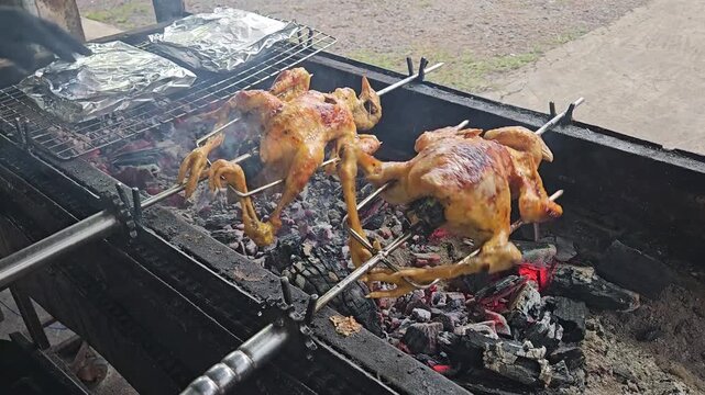 Two whole marinated chickens being roasted on a rotisserie over charcoal at a Vietnamese street food stall, with smoke and glowing embers.