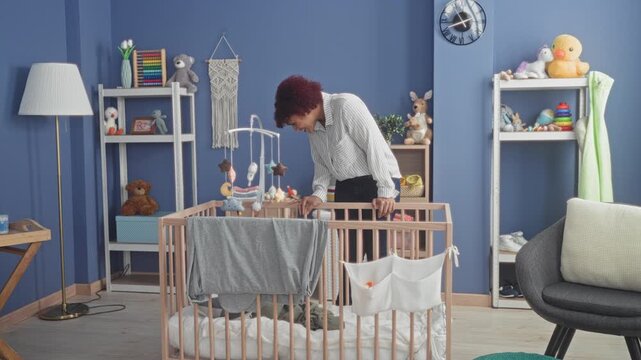 Woman leaning over crib touching a hanging mobile in a studio nursery, smiling while checking the bassinet; tenderness bonding.