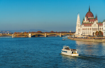 Naklejka premium Vibrant cityscape view of Budapest's magnificent Parliament building along the Danube River featuring a tourist boat under the iconic Hungarian bridge architecture travel destination