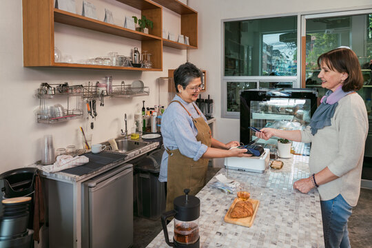 Wide shot of a friendly Asian barista processing a contactless payment for a senior customer in her cozy, local coffee shop. Concept of small business, entrepreneurship, and daily operations.