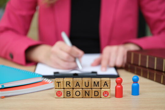 Brightly colored books and wooden letters spell out trauma bond. A person in a pink blazer writes in a notebook, symbolizing the journey of healing and self-discovery.