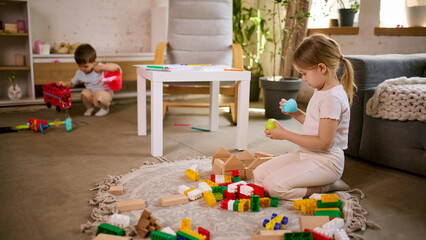 Fototapeta premium Little girl playing with toys while boy in background making mess in living room. Concept of parenting reality content, childcare challenges, home routine situations and family lifestyle storytelling.