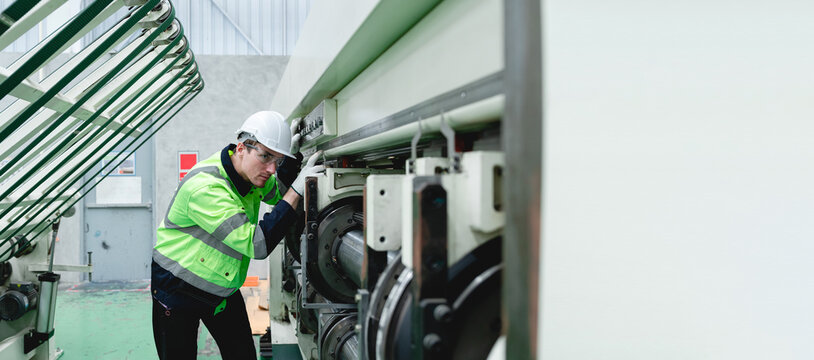 Man in a white helmet and bright green workwear inspects complex machinery within a gray industrial factory setting, wearing dark pants and sneakers, performing a detailed maintenance inspection.