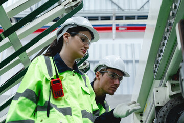 Two workers, a man and a woman, conduct a detailed safety inspection of a green corrugated cardboard pipe with a handheld device while wearing hard hats, safety glasses, and high-visibility yellow jac © AU USAnakul