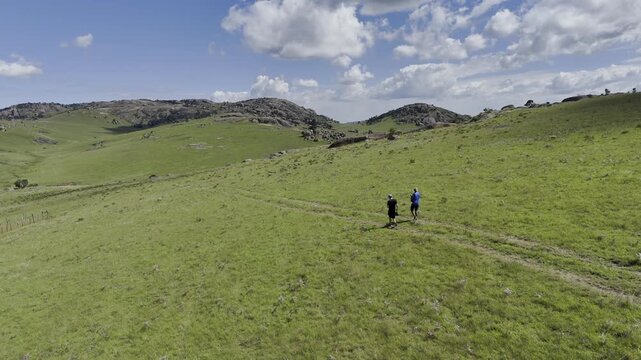 Drone hovers over green field with rocky hills as two hikers walk through on a sunny day at Sibebe Rock near Mbabane, Eswatini