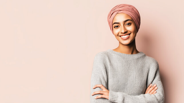 Cheerful young woman expressing happiness, wearing a pink headscarf and a gray sweater, standing with arms crossed