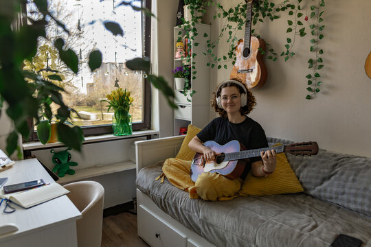 Teenager with curly hair playing guitar in cozy bedroom