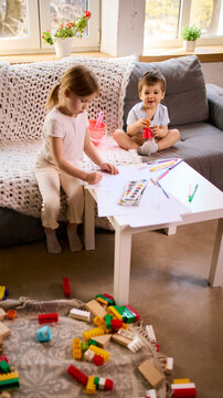 Little children sitting on sofa drawing with pencils and playing with toys in bright living room. Concept of kids lifestyle content, creative play products promo and family home environment visuals.