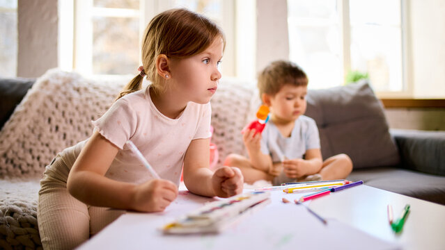 Little girl focused on drawing at table while boy sitting behind playing with toy at home. Concept of concentration during tasks, background distraction scenarios and learning environment at home.