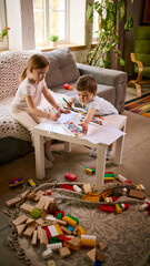 Fototapeta premium Little children sitting at small table drawing with pencils and watercolor paints at home. Concept of preschool education materials, creative learning tools and kids activity product promotion.
