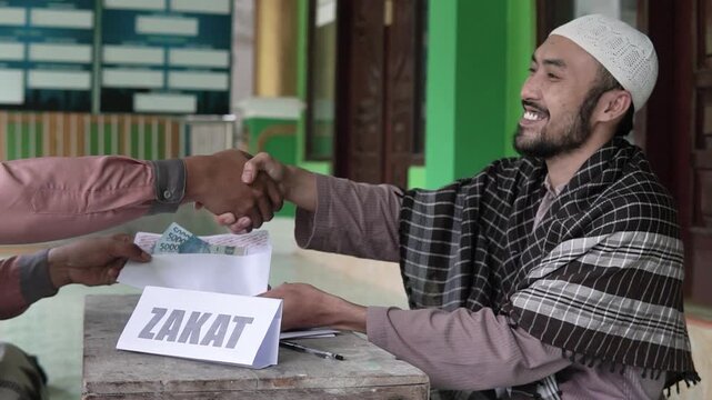 Zakat donation handover at mosque table, man passing envelope of cash to recipient with visible ZAKAT sign, warm handshake