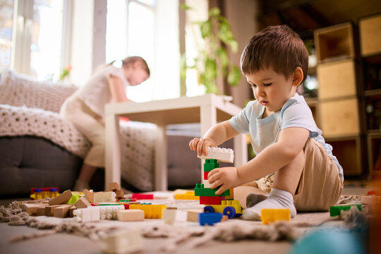 Little boy assembling colorful block structure while girl drawing at table in background at home. Concept of multitasking parenting visuals, kids activities diversity and home learning environment.