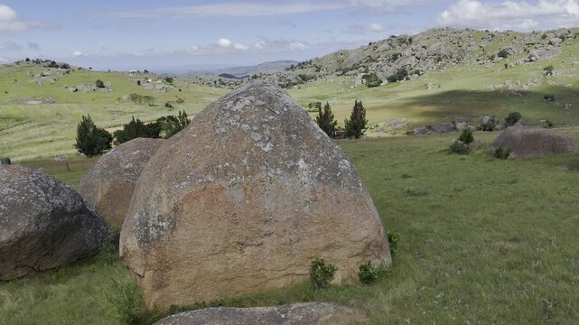 Drone slowly flies to right, following two hikers walking through grassy field with large boulders on a sunny day at Sibebe Rock near Mbabane, Eswatini