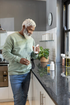 Senior African American man leaning holding white pill bottle and water glass in modern kitchen