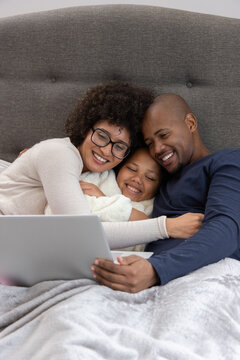 Diverse family cuddling on bed in grey blanket, father holding laptop, mother wearing glasses