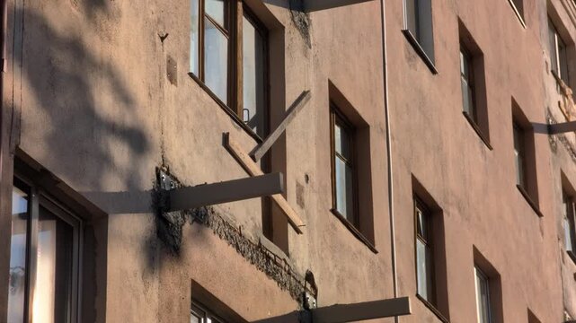 Multi-story residential building facade undergoing renovation. Demolished balconies expose concrete, metal beams. Sunlight casts shadows on uneven wall. Windows reflect blue sky.