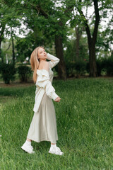 Fototapeta premium A stylish, beautiful young woman in a light dress poses in a green park. Spring fashion, a natural look, and a relaxed mood.