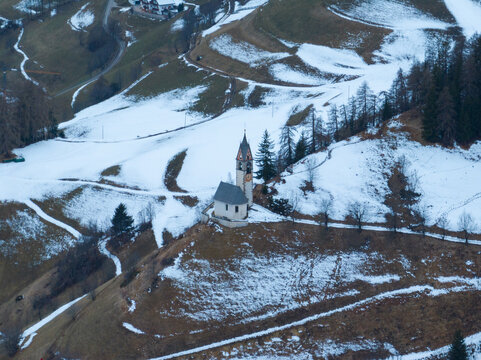 Aerial view of the Cappellina di Santa Barbara chapel perched on a snowy hillside with pine trees and winding mountain paths in Morter, Trentino-Alto Adige, Italy.