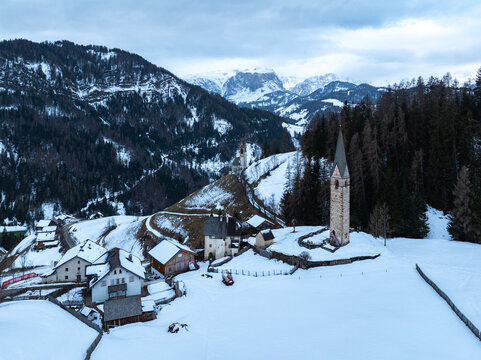 Aerial view of Cappellina di Santa Barbara and traditional houses in a snowy mountain landscape with pine forests and peaks in Morter, Trentino-Alto Adige, Italy.