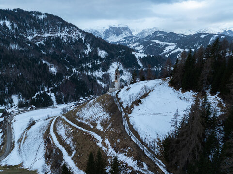 Aerial view of Cappellina di Santa Barbara on a snowy hillside surrounded by dense pine forests and majestic snow-capped mountains Morter, Trentino-Alto Adige, Italy.