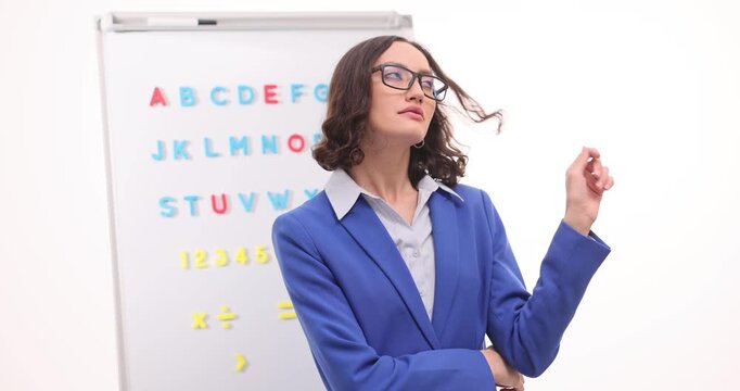 Lady teacher in glasses stands near whiteboard gently twisting strand of hair around finger in white classroom. Female crosses arms. Slow motion