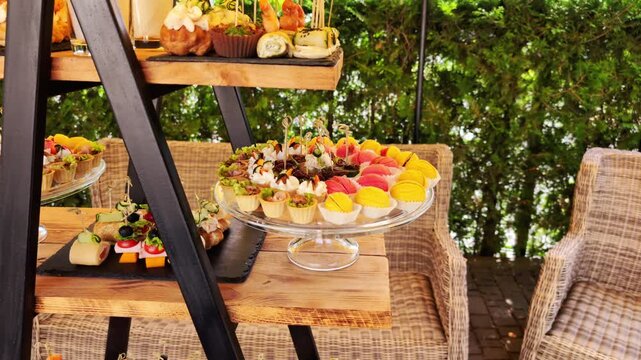 Outdoor catering display presents diverse gourmet appetizers, desserts. Colorful macarons, savory tartlets, canap s, finger foods on tiered wooden stand, ready for event guests