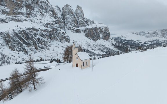 Aerial view of Cappella di San Maurizio surrounded by deep snow and towering rocky mountain peaks in Selva di Val Gardena, Trentino-Alto Adige, Italy.