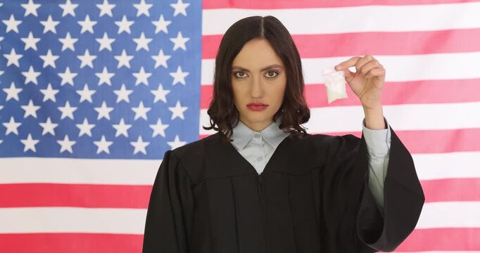Serious lady judge in black judicial robe stands against American flag. Woman holds small plastic bag of drugs and pair of handcuffs. Slow motion