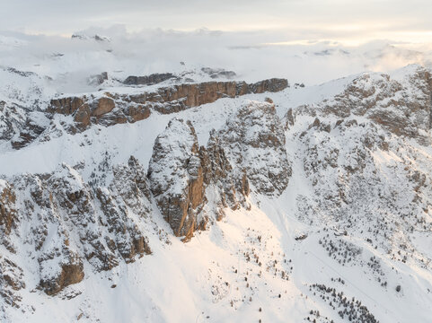 Aerial view of snow-covered jagged mountain peaks and rocky cliffs under a misty sky with soft golden light in Selva di Val Gardena, Trentino-Alto Adige, Italy.