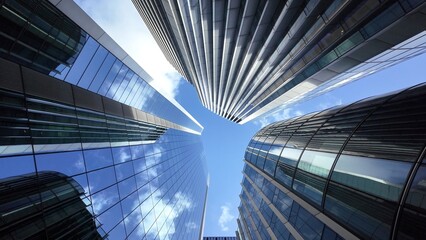 Looking up at modern glass and steel skyscrapers reflecting a clear blue sky with white clouds, capturing themes of business, finance, and urban development in a bustling city © DawDunia
