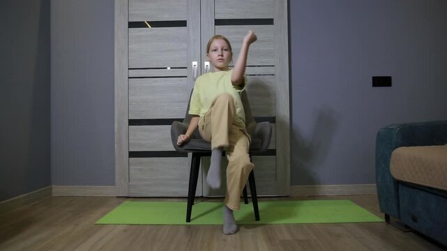 Child performs seated leg lifts and arm movements on chair in apartment, demonstrating simple indoor workout, promoting active lifestyle and well-being