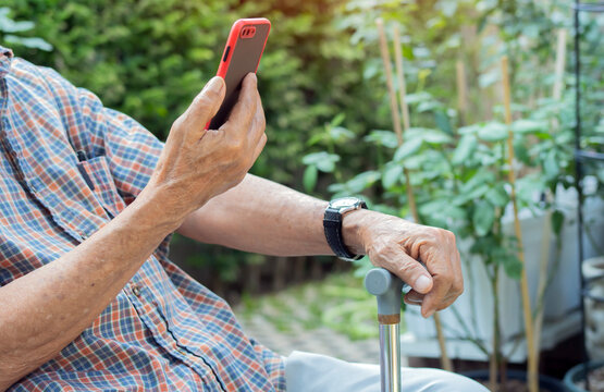 Side view of unrecognizable elderly man sitting at park with walking stick and using his smart phone