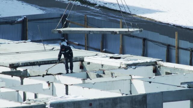 Construction worker clears snow and debris from partially built concrete frame of building on cold winter day. Large concrete slab suspended by chains from crane descends above structure, showing ongo