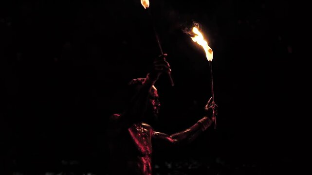 Male performer engages in intense night show, holding two burning torches against black background, firelight illuminating body outline, showcasing ancient art of fire manipulation.