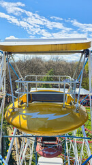  close-up of empty yellow ferris wheel gondola with white canopy roof and metal safety rail, white blue steel structure around, trees and blue sky with clouds in background © Татьяна Швец