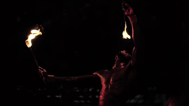 Close up shot of shirtless man performing thrilling fire act at night, holding one flaming torch and balancing another with fire in mouth, dramatic display of fiery art and extreme showmanship