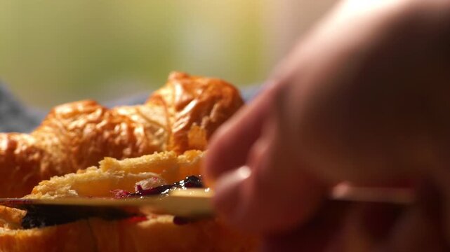 Close-up of a knife spreading glossy fruit jam onto a flaky croissant. Rich textures and warm tones highlight a cozy breakfast moment with fresh pastry and delicious topping in soft natural light.