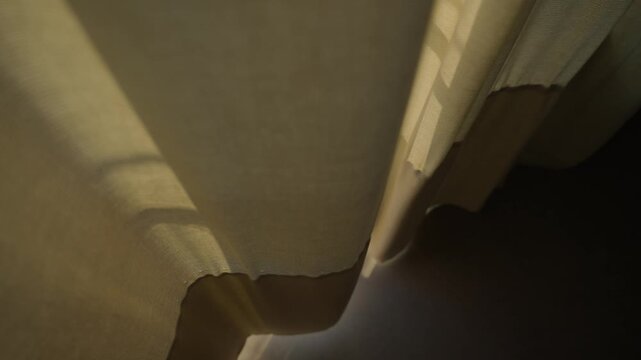Window curtain moving slowly by wind letting sunlight shine inside house room. Dark indoor apartment or home as sun is entering by small crack made by yellow window fabric drape blowing in wind