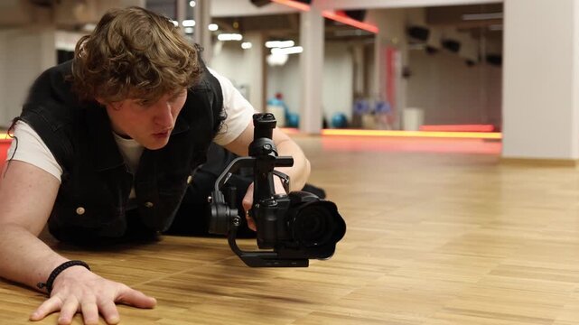Videographer shoots low angle scene lying on floor using camera on stabilizer focusing on creative framing perspective control and smooth motion in indoor studio during professional filming practice