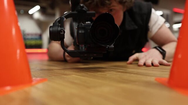 Videographer lies on floor holding camera on stabilizer practicing low angle shot focusing on framing precision movement control and creative perspective in modern indoor studio training session