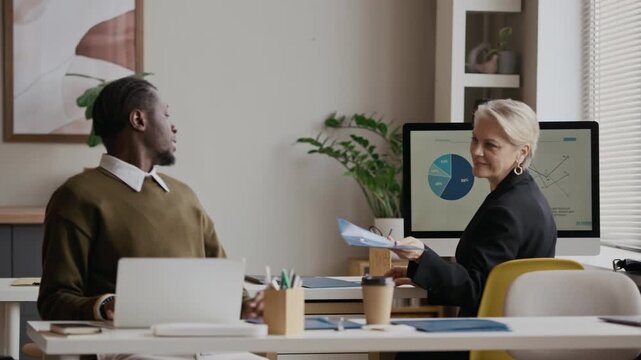 Medium shot of senior Caucasian woman handing file folder with documents to young Black male colleague while working in modern open space office