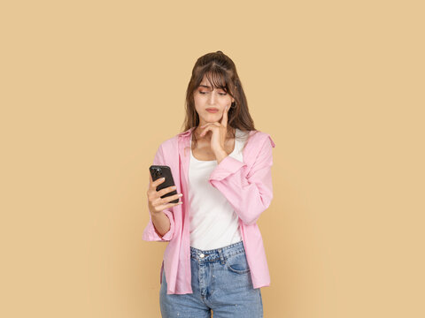 Doubtful young woman holding smartphone and touching chin with skeptical expression, evaluating message or decision concept. Isolated on beige background with copy space for advertising use.